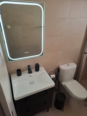 A bathroom with a white sink, a mirror with a light, a toilet, and a black trash bin. - Bright and comfortable apartment for a pleasant stay (Βάρη)