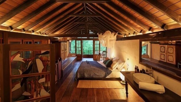 A room with a sloped ceiling, wooden flooring, a bed, a bench, a large window, and a mirror. - Sama Retreats (Galle)