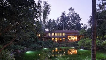 Um edifício de dois andares com um lago espelhado na frente, cercado por vegetação tropical exuberante.