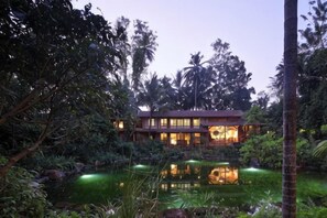 A two-story building with a reflective pond in front, surrounded by dense tropical vegetation.