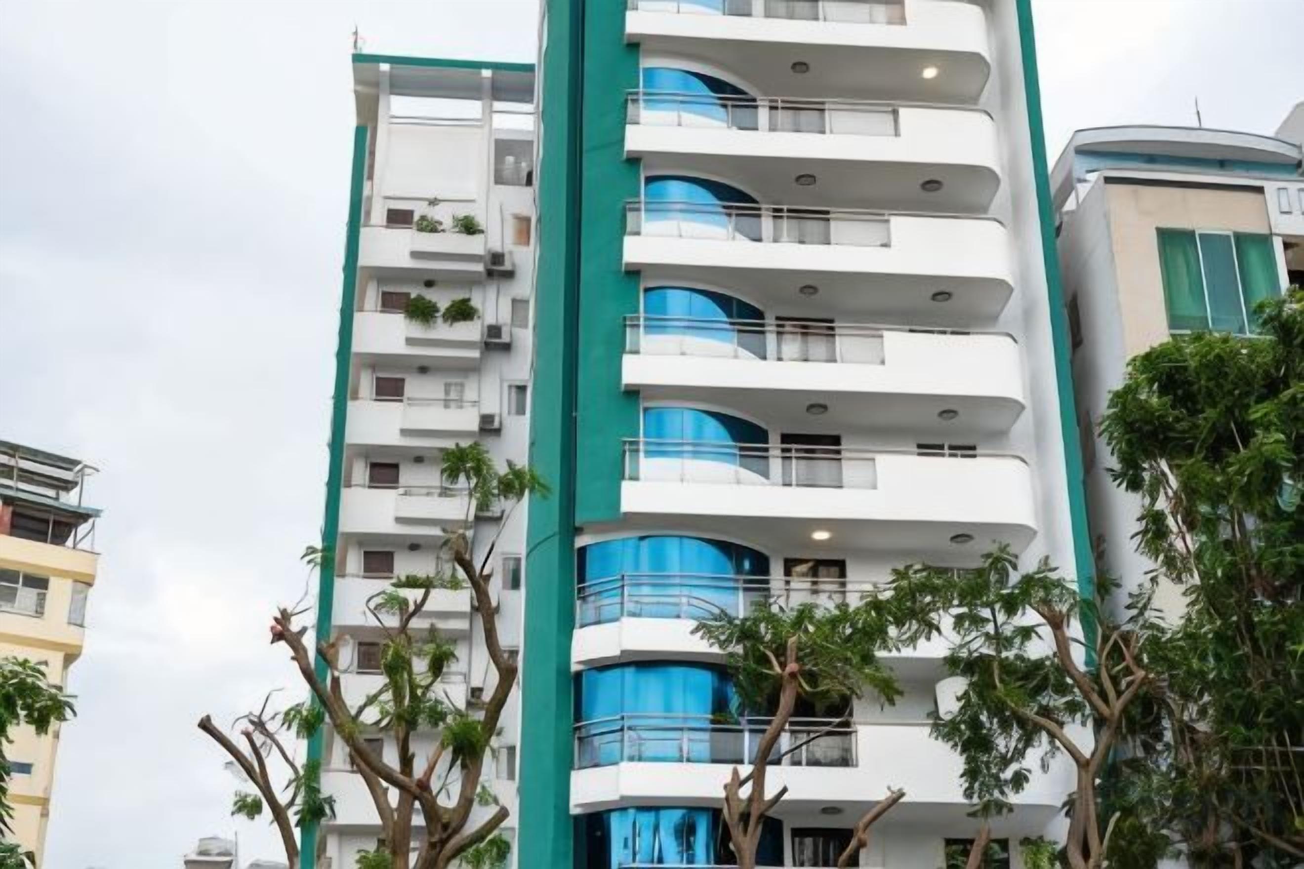 Um edifício de vários andares com varandas, um pilar central verde e árvores ao redor.