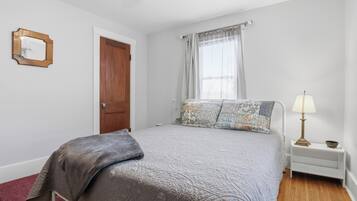 Quarto com cama, mesa de cabeceira, luminária, porta e janela com cortinas.