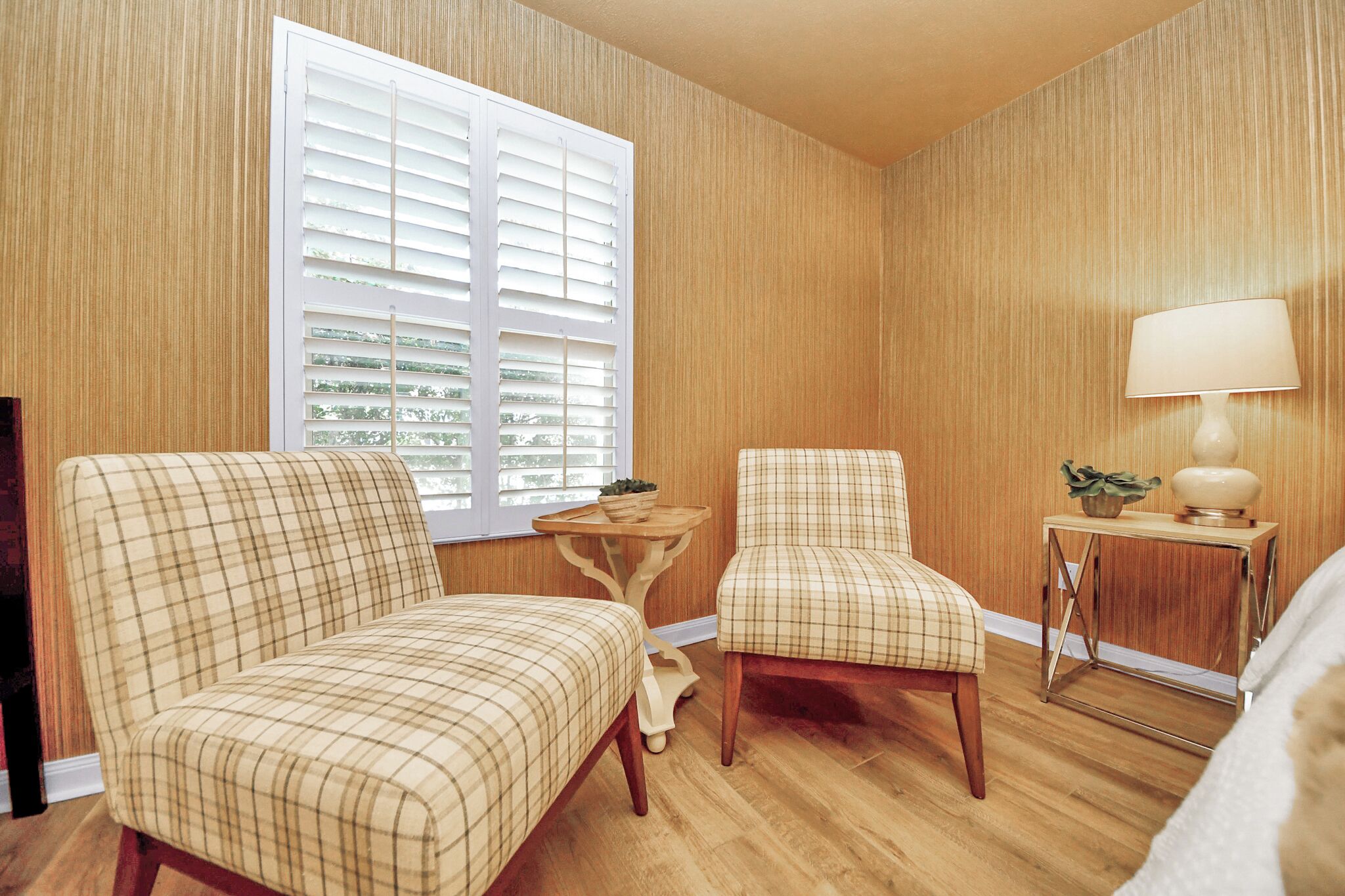 A room with two plaid chairs, a small wooden table with a lamp, and a window with blinds.