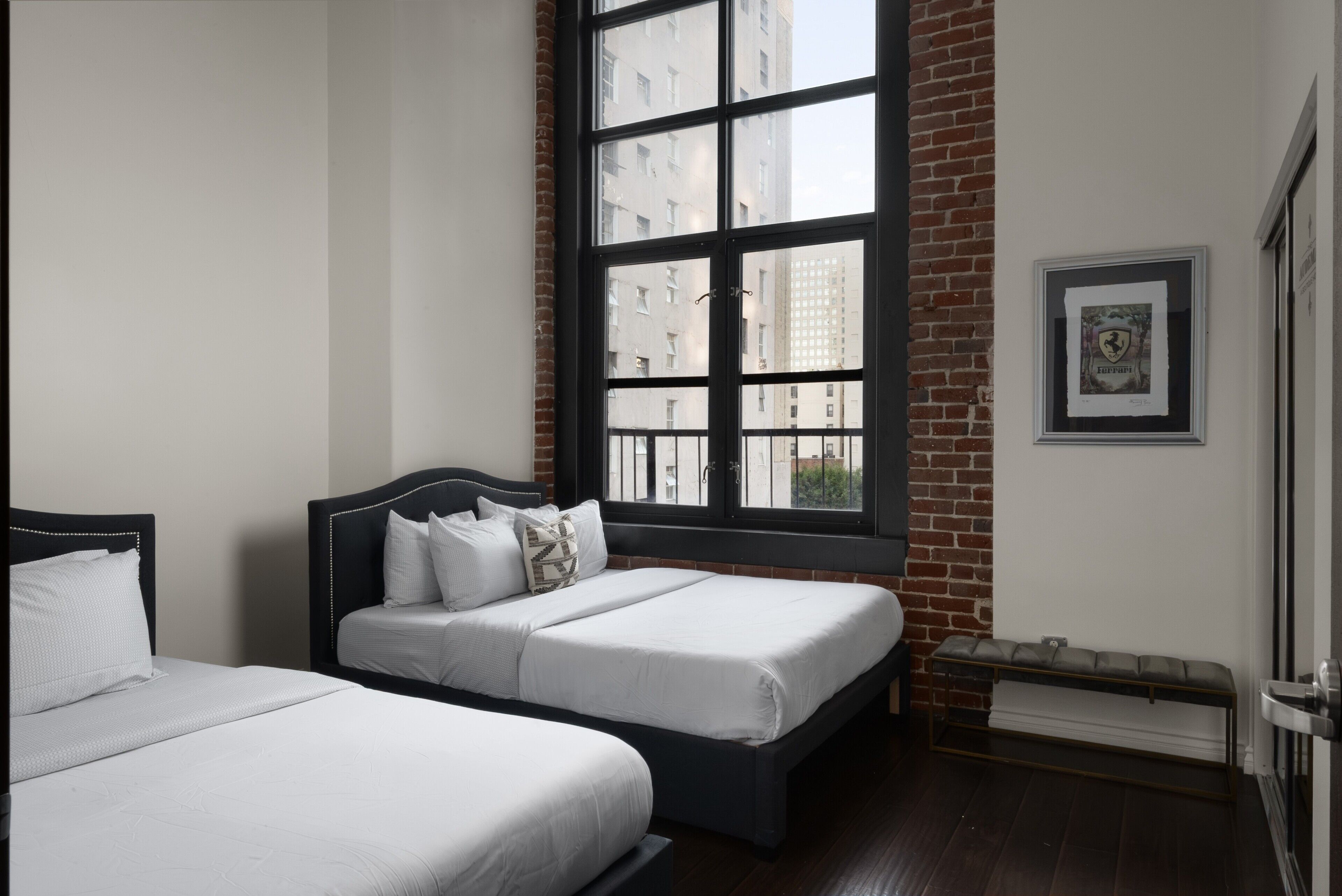 A hotel room with two beds, a large window with a view of buildings, a framed picture on the wall, and a bench by the window.
