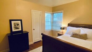Une chambre à coucher avec un lit, une table de chevet, une photo encadrée, une porte et une fenêtre avec des stores.