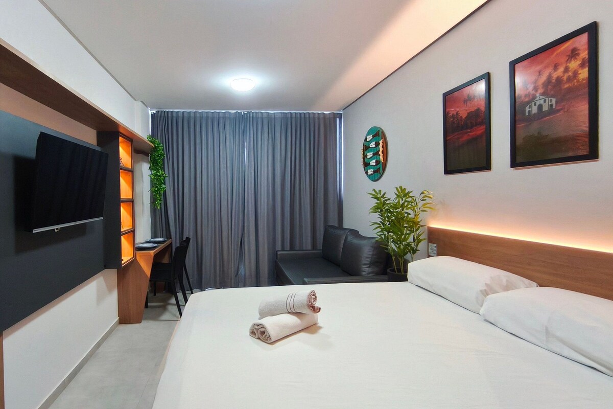 A hotel room with a bed, a TV, a desk, a chair, a sofa, a plant, and artwork on the walls.