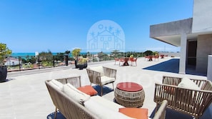 Une terrasse sur le toit, meublée de mobilier en osier, avec vue sur l’océan et un bâtiment en arrière-plan.