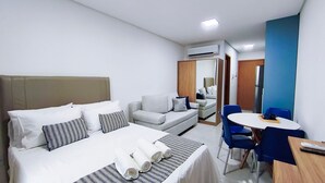 A hotel room with a bed, a sofa, a dining table with chairs, an air conditioning unit, and a mirror. - Acqua Beach Residence (Tamandaré)