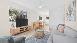 Ein modernes Wohnzimmer mit Flachbildfernseher, grauem Sofa, Couchtisch aus Weidengeflecht und einem Essbereich mit Tisch und Stühlen.