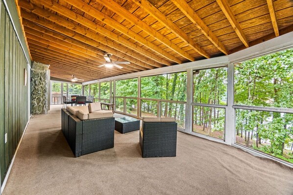 A covered outdoor seating area with a ceiling fan, large windows, and a view of trees.