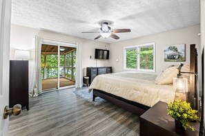 Un dormitorio con cama, ventilador de techo, televisión y ventana con vistas a la vegetación.
