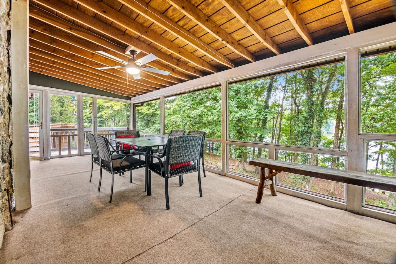 Un patio couvert avec une table et des chaises, de grandes fenêtres donnant sur la verdure et un ventilateur de plafond.