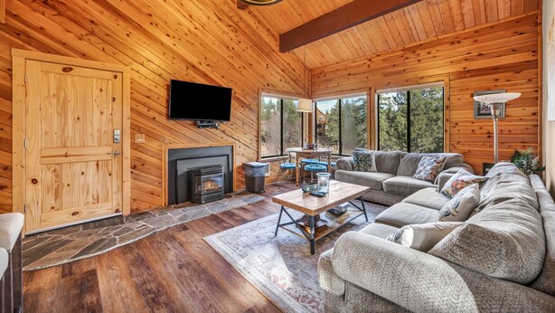 A wooden cabin interior with a fireplace, a television, a sofa, and large windows offering a view of the forest. - Tahoe Donner Getaway | Ski-In/Ski-Out Retreat (Truckee)