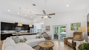 A spacious living area with a white sofa, a round wooden coffee table, and a wicker chair. A kitchen with dark cabinets and a countertop is visible in the background. - Sunshine and Serenity Holidays (Boynton Beach)