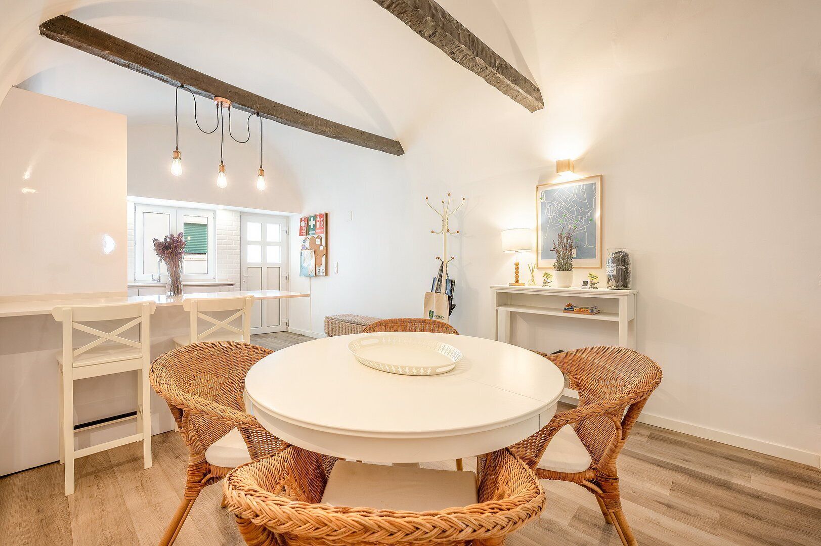 Een eethoek met een ronde tafel, rieten stoelen en een witte tafelkleed. Er staat een witte kast met een kaart en een lamp erop.