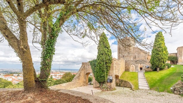 A historic stone building with a paved pathway leading to an entrance, surrounded by trees and a view of a distant cityscape.