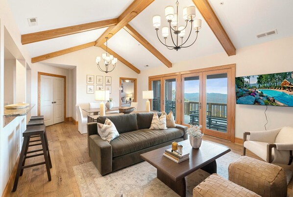 A spacious living room with a high ceiling, a dark brown sofa, a coffee table, and a large window offering a view of a mountain landscape. - Grand Oaks RQ6- Mountain Condo, Gated Resort, Indoor/Outdoor Pools & Hot Tub (Banner Elk)
