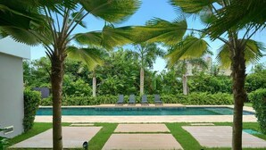Zona de piscina con palmeras, cielo despejado y un camino que lleva a sillones de descanso.
