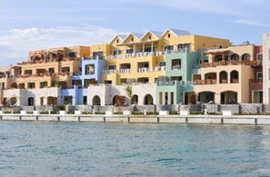 A row of colorful hotel buildings with balconies, a stone wall in front, and a body of water in the foreground. - New! Modern & Stylish Cap Cana Retreat with Pool (Punta Cana)