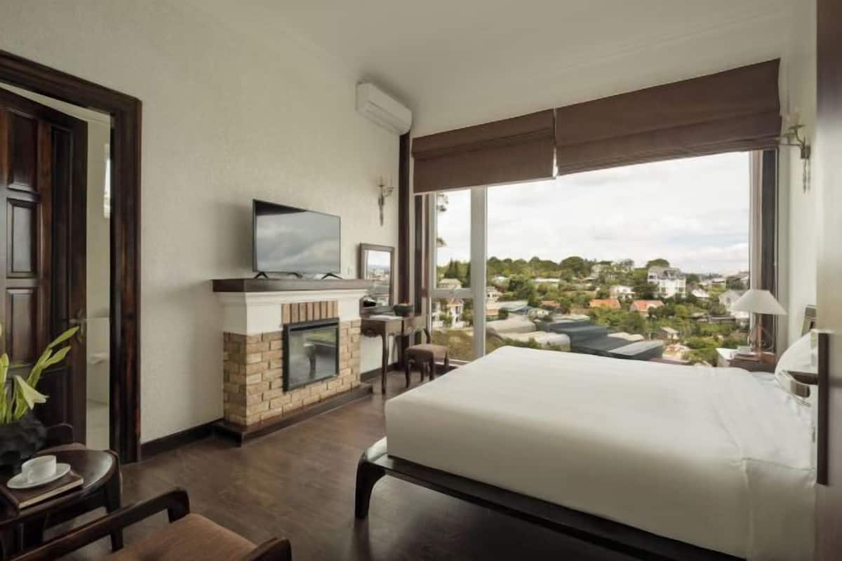 A hotel room with a bed, a television, a fireplace, a dining table, and a view of the city.