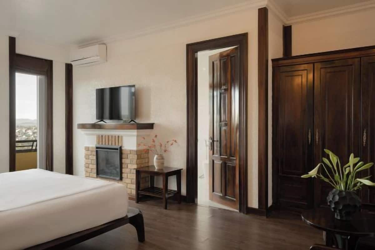 A bedroom with a bed, a TV mounted above a fireplace, a door leading to another room, and a view through a window.