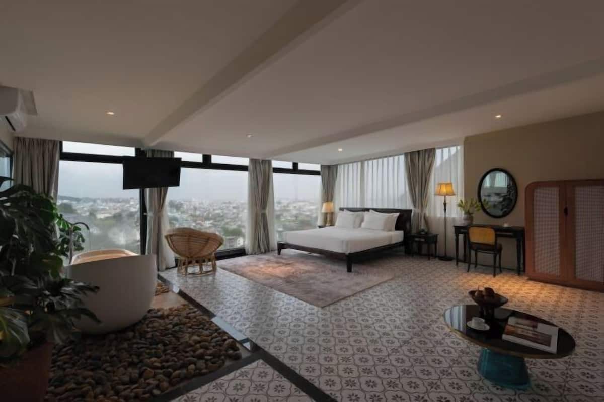 A hotel room with a large bed, a TV, a desk with a lamp, and a view of the city.