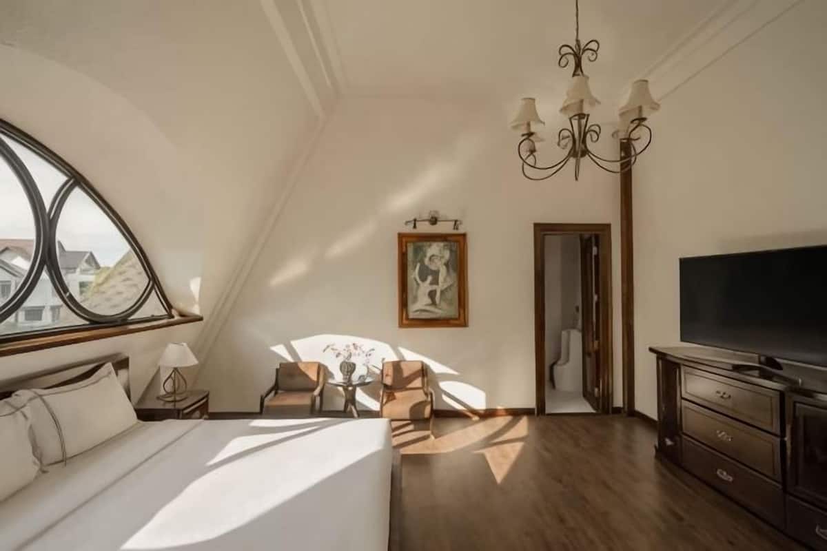 A bedroom with a bed, a TV, a chandelier, a painting, and a window with a view.
