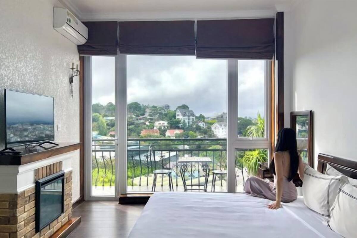 A hotel room with a large bed, a TV, a fireplace, and a balcony with a view of the city.