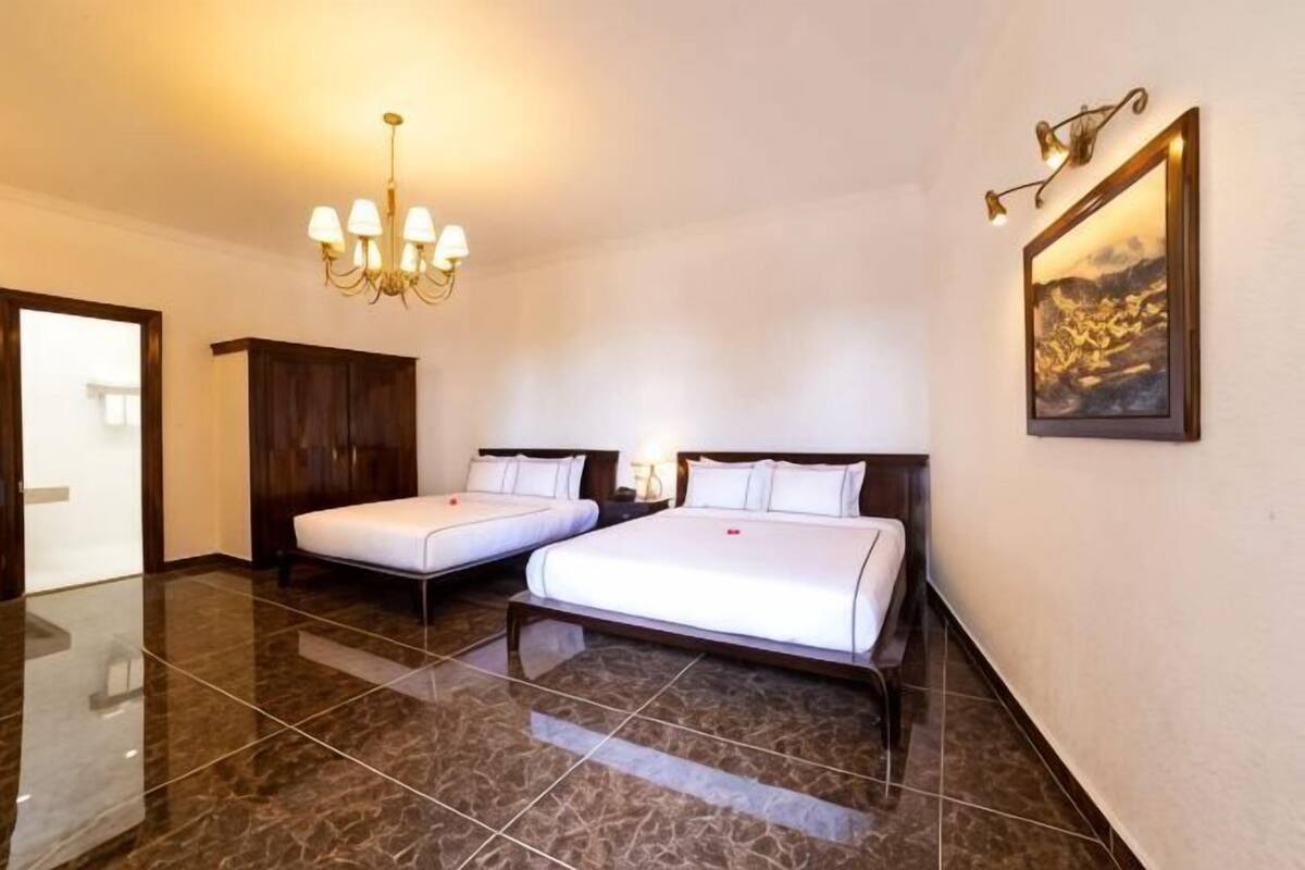 A hotel room with two beds, a chandelier, a painting, and a wooden wardrobe.