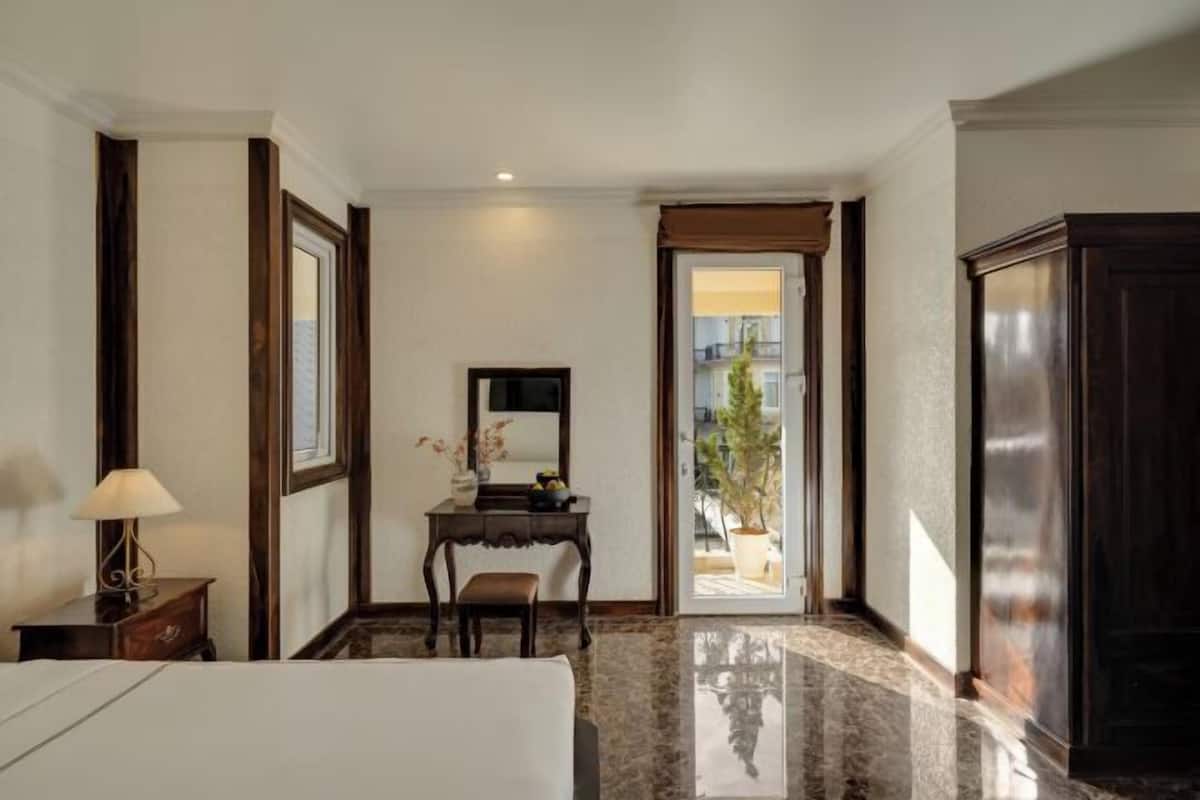 A hotel room with a bed, a desk with a chair, a lamp, a mirror, and a view of the outside.