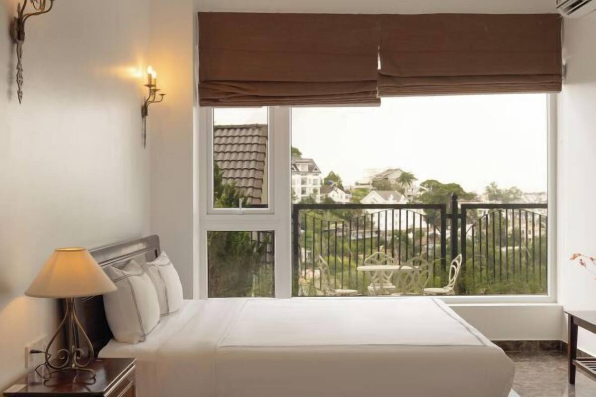 A hotel room with a bed, a nightstand with a lamp, a window with a view of the outside, and a balcony with a fence.