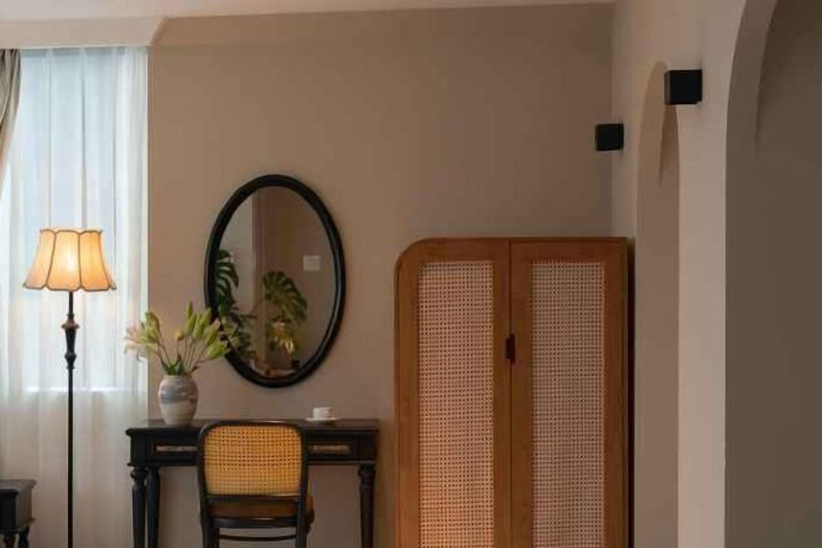 A room with a desk, chair, lamp, mirror, and a wooden wardrobe.