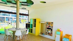A modern, well-lit room with a large, light blue sectional sofa, a round table with chairs, and a colorful play area with toys and books. - Makia Beach Experience Resort - Muro Alto, Porto de Galinhas - B18 POOL FRONT (Ipojuca)