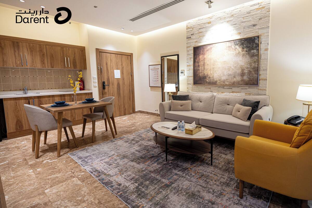 Ein modernes Wohnzimmer mit einer Couch, einem Couchtisch und einem gelben Sessel. Es gibt einen Essbereich mit einem Holztisch und Stühlen. Die Wand des Raumes ist aus Stein und über der Couch hängt ein Gemälde.