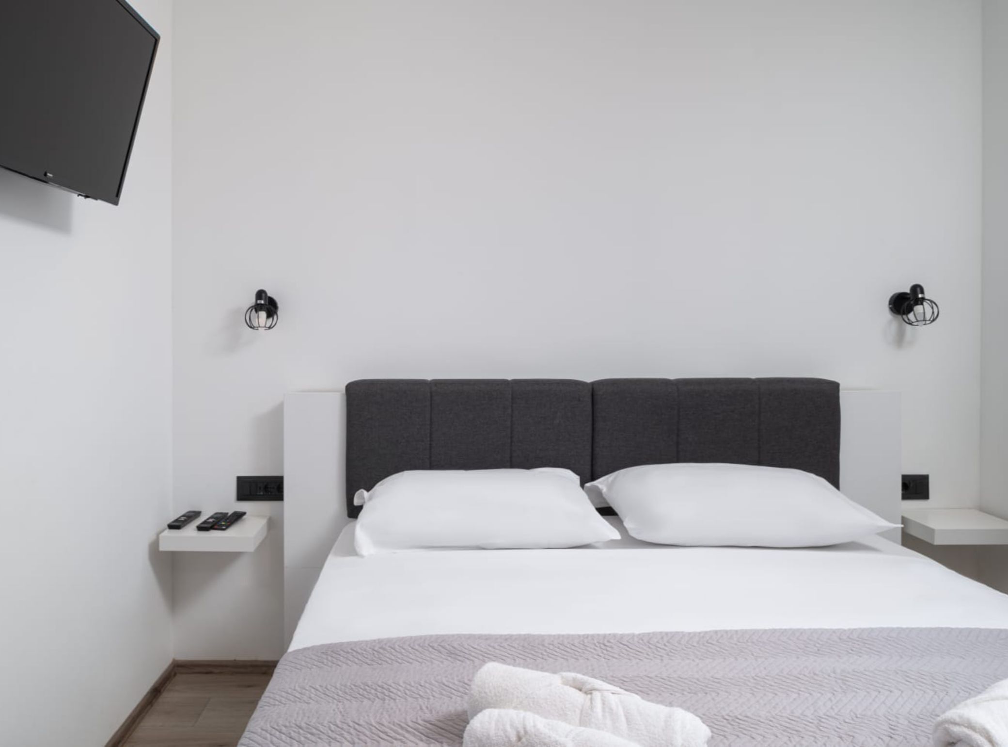 Una habitación de hotel moderna con una cama grande, ropa de cama blanca, un cabecero gris, dos mesitas de noche con lámparas y un televisor montado.