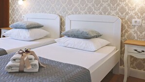 Une chambre d’hôtel avec deux lits, chacun agrémenté de draps blancs et d’oreillers gris. On y trouve des tables de chevet avec des lampes et une petite table de nuit en bois. Les murs sont recouverts d’un papier peint à motifs.