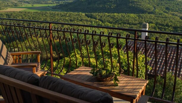 A balcony with a wooden table and chairs, overlooking a lush green landscape with hills and a distant building. - Istra Green Soul Apartment (Oprtalj)