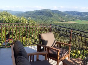 A balcony with a wooden chair and a sofa, overlooking a vineyard and distant hills. - Istra Green Soul Apartment (Oprtalj)