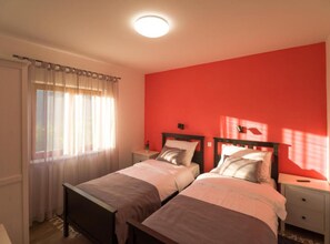 A bedroom with two beds, a red wall, a window with sheer curtains, and a ceiling light. - Istra Green Soul Apartment (Oprtalj)