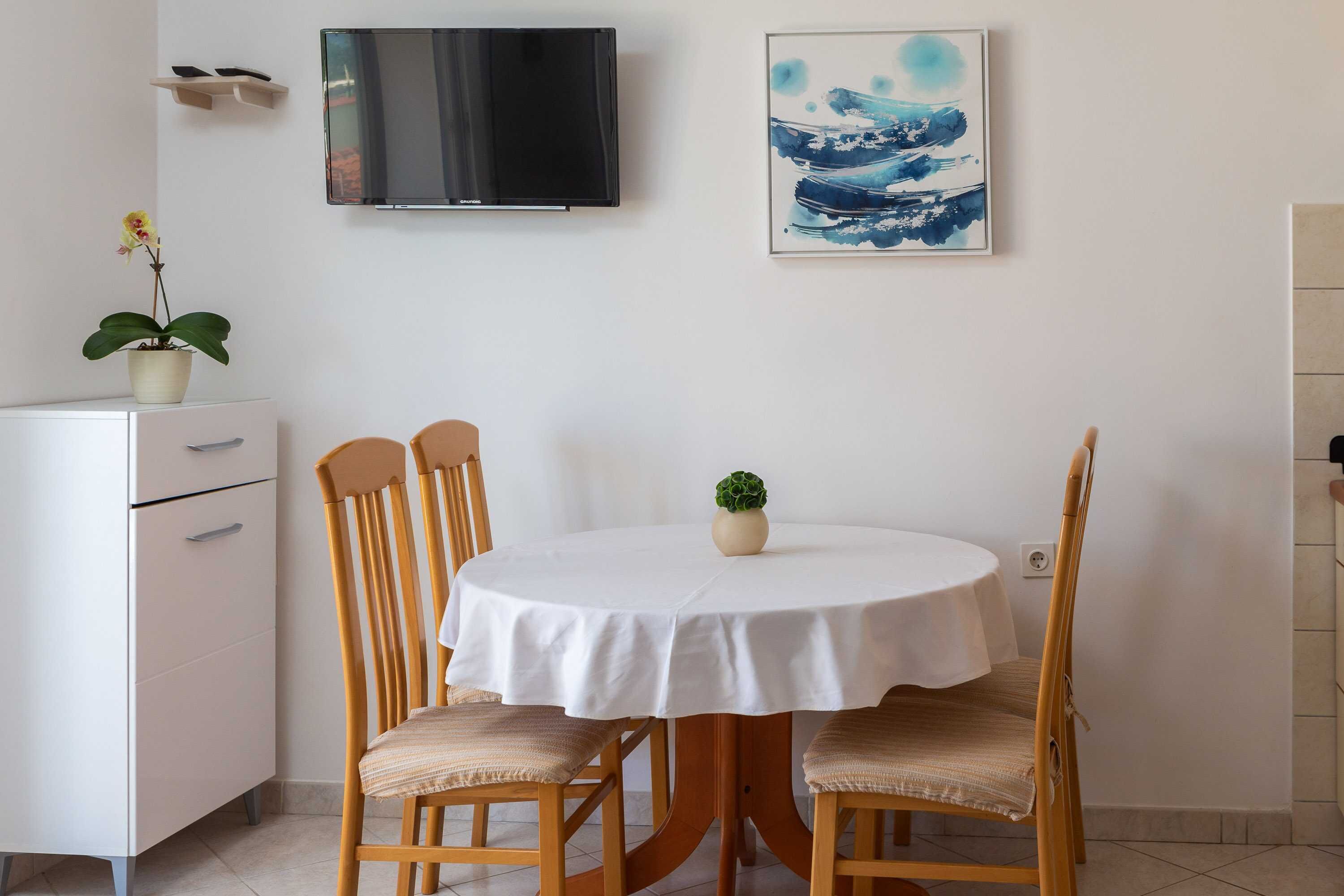Un comedor con una mesa redonda, cuatro sillas de madera, un armario blanco, un televisor montado en la pared y una obra de arte enmarcada que representa una ola.