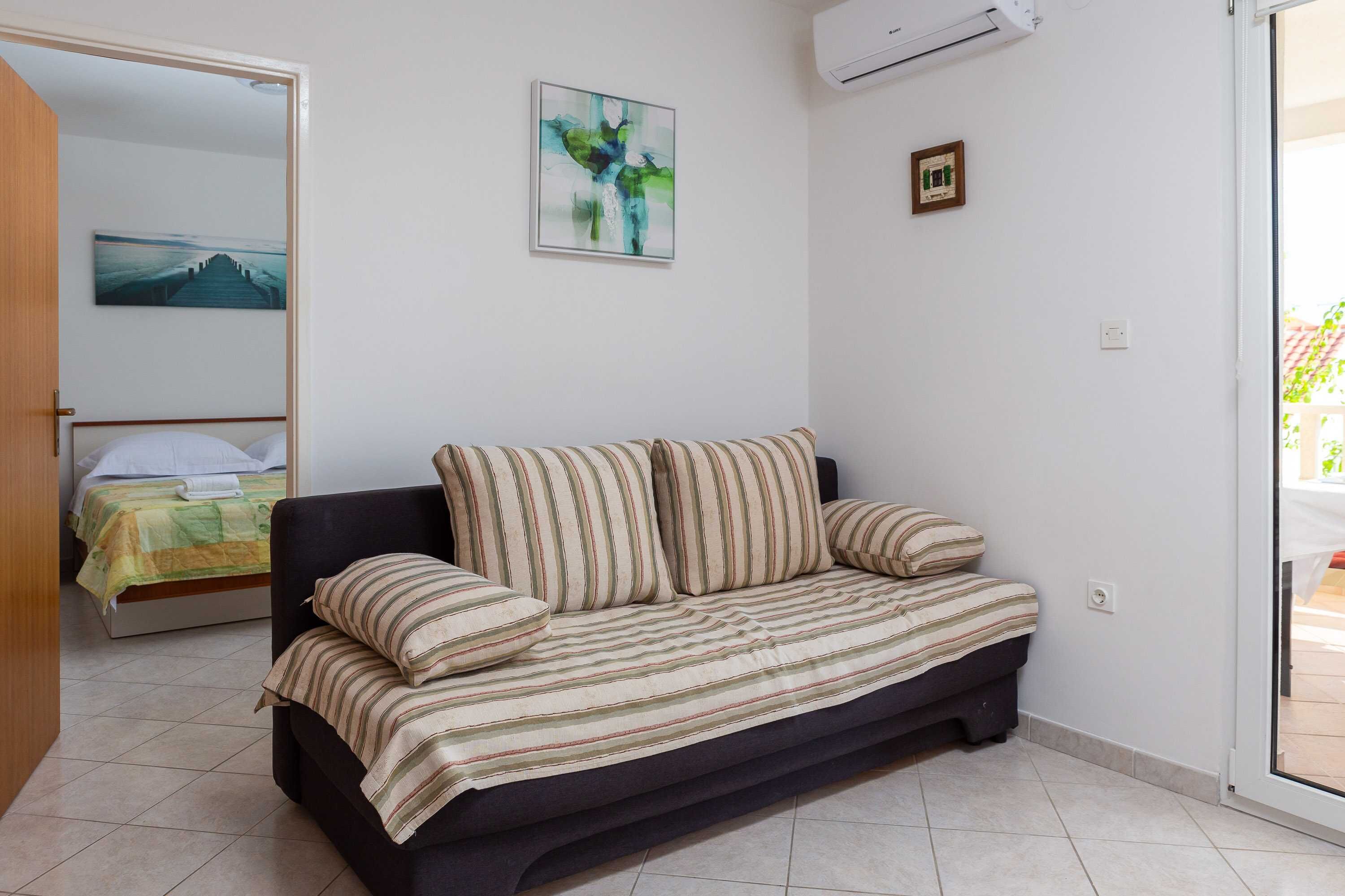 Una habitación con un sofá oscuro, una cama con colcha a rayas verde y amarilla, un cuadro en la pared y un equipo de aire acondicionado.