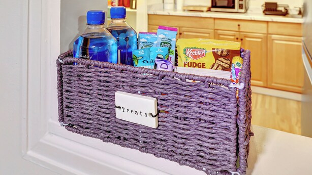 Ein Korbflechtschrank mit dem Aufdruck „Snacks“ ist mit verschiedenen Getränken und Speisen gefüllt und auf einer weißen Tür platziert.