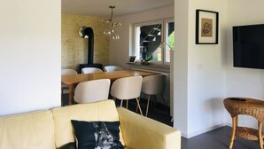 Ein modernes Wohnzimmer mit einem gelben Sofa, einem Essbereich mit Holztisch und -stühlen, einem schwarzen Kamin und einem großen Fenster mit Blick auf eine Treppe.
