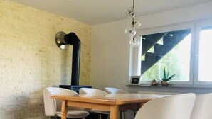 Ein Essbereich mit Holztisch und weißen Stühlen, einer Ziegelwand, einer schwarzen Rohrleitung, einer Pendelleuchte und einem Fenster mit Blick auf Grünflächen.