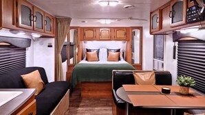 A compact RV interior with a bed, a sofa, a dining table, and a kitchenette.