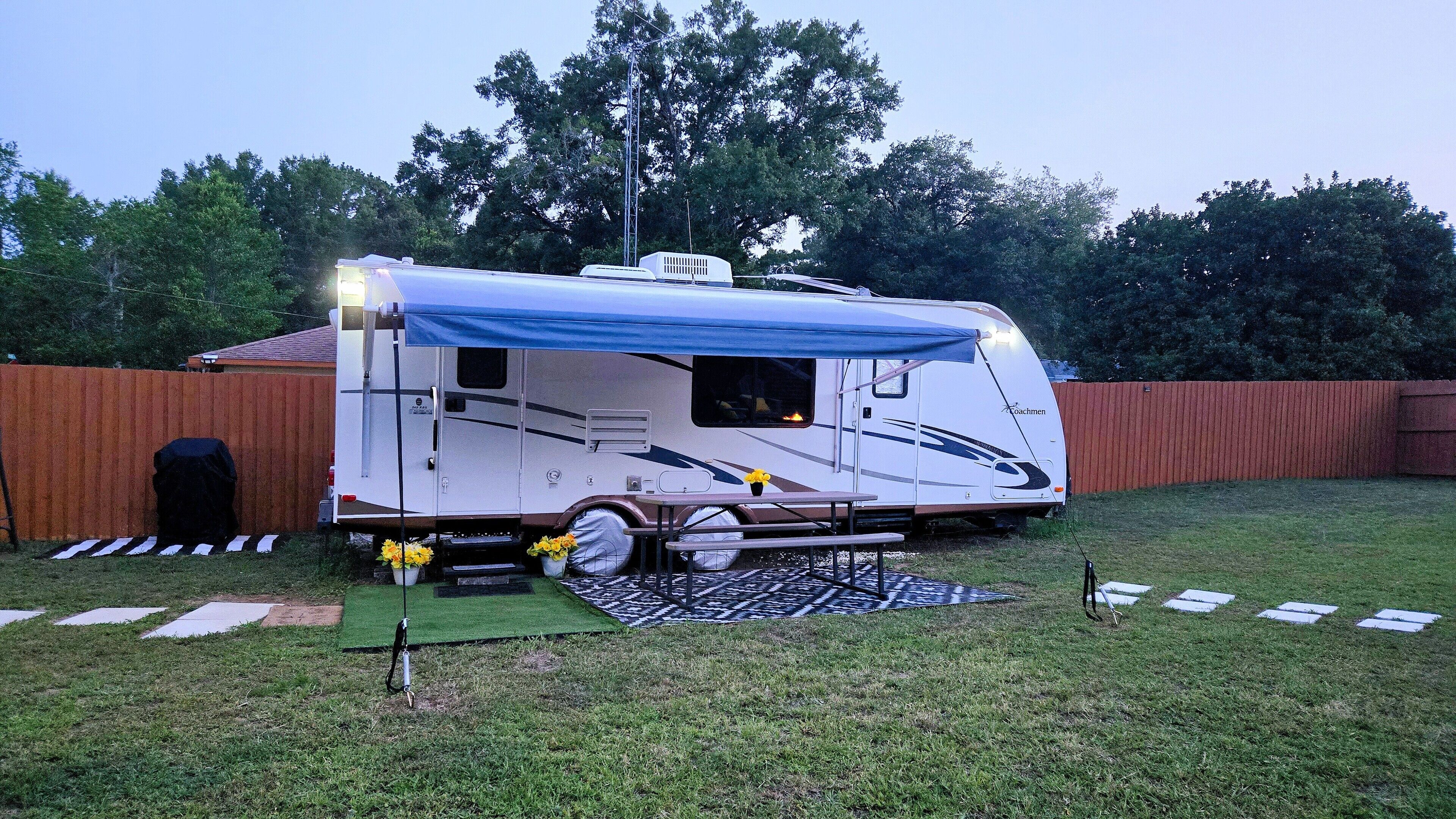 Un camper parcheggiato in un giardino sul retro, con una tenda blu, un tavolino e vasi di piante.