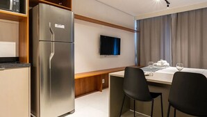 A modern kitchen with a built-in refrigerator, a microwave, and a flat-screen TV on the wall. There is a dining area with a table, chairs, and a wine glass. - Movi Life Torre 2 (São Paulo)