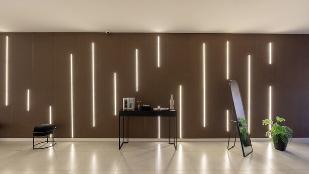 A modern interior with a dark brown wall, a black table with books and a vase, a black stool, and a plant. - Movi Life Torre 2 (São Paulo)
