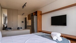 A hotel room with a bed, a flat-screen TV mounted on the wall, a wooden headboard, a small desk with a chair, and a kitchenette area. - Movi Life Torre 2 (São Paulo)
