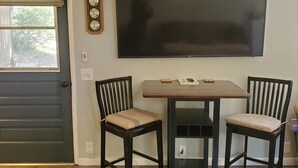 Een eetgedeelte met een tafel en twee stoelen, een flatscreen televisie aan de muur, een raam met uitzicht op bomen en een deur.
